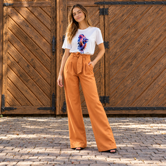 A woman stands outdoors in front of large rustic wooden barn-style doors, wearing a white graphic T-shirt and high-waisted wide-leg orange pants with a tie waist.