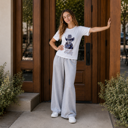 A woman stands in front of a wooden door wearing a white graphic T-shirt with a black cowboy illustration and the words 'WANTED COWBOY' and light gray wide-leg sweatpants.